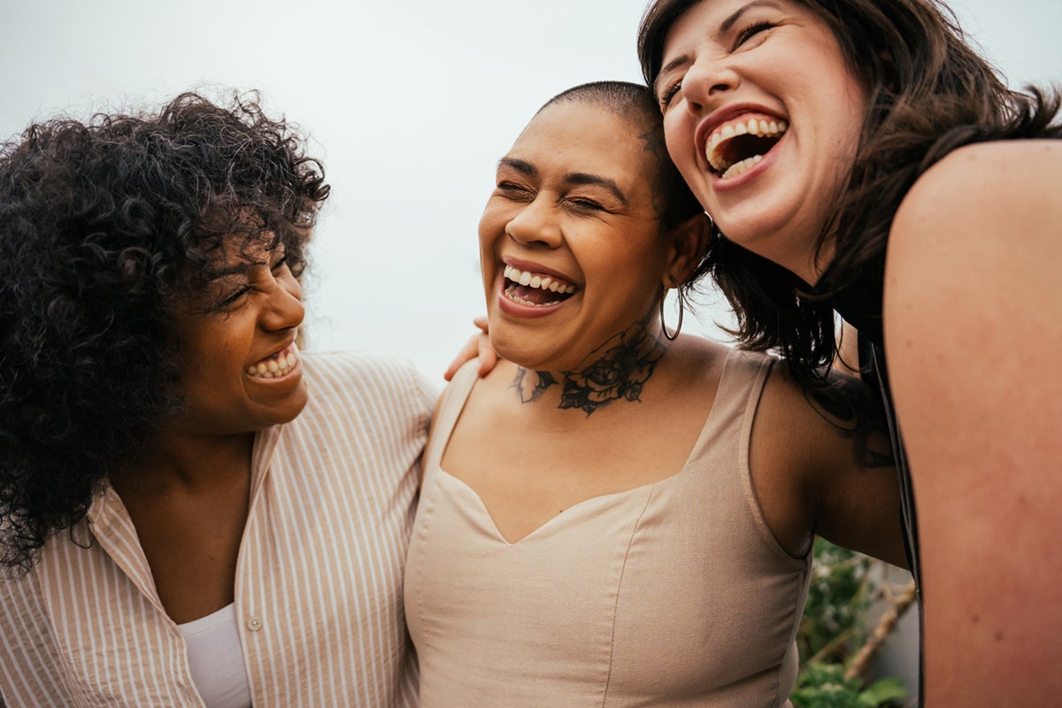 Three women standing close together, smiling and laughing joyfully outdoors.