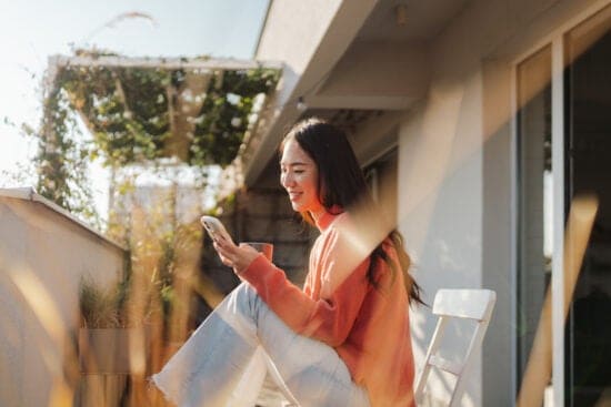 Woman in an orange sweater sits on a balcony, smiling at her phone in sunlight, unaware of third party cookies tracking her browsing.