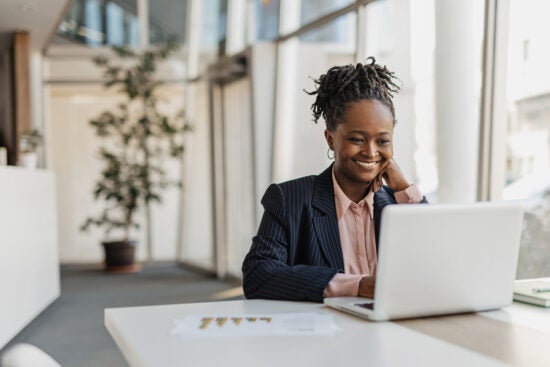 Smiling woman in a suit working on a laptop at a desk in a bright, modern office.