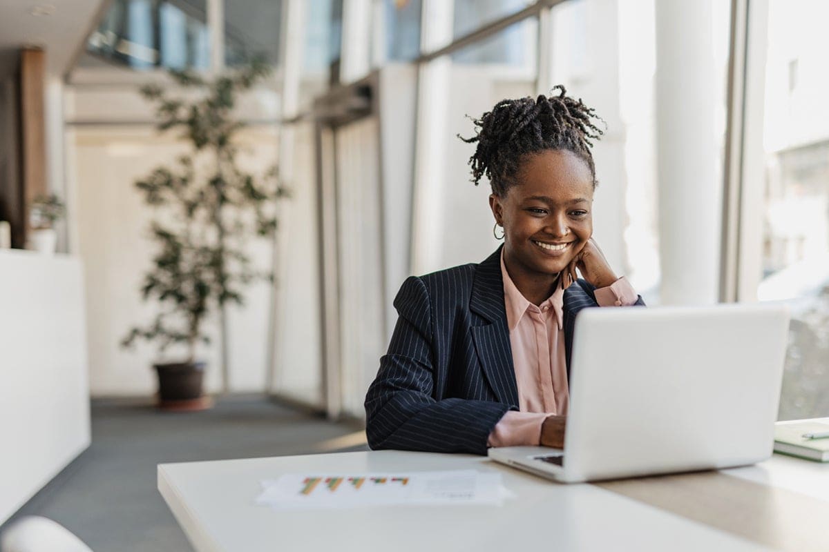 Smiling woman in a suit working on a laptop at a desk in a bright, modern office.