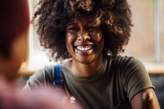 A person with curly hair smiles warmly, seated indoors near a window, wearing a green shirt.