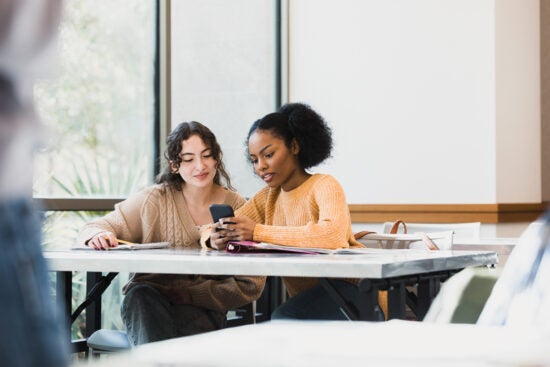 Two women sit at a table, looking at a smartphone together, with notebooks and papers in front of them.