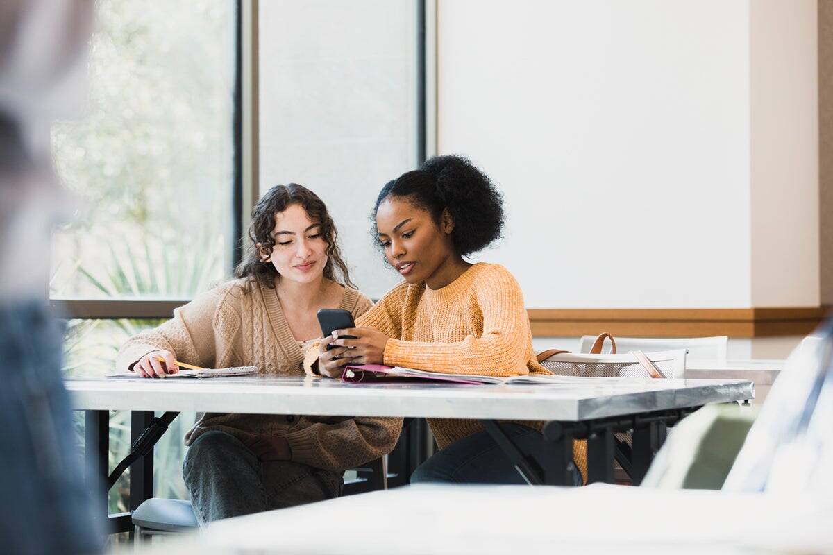 Two women sit at a table, looking at a smartphone together, with notebooks and papers in front of them.