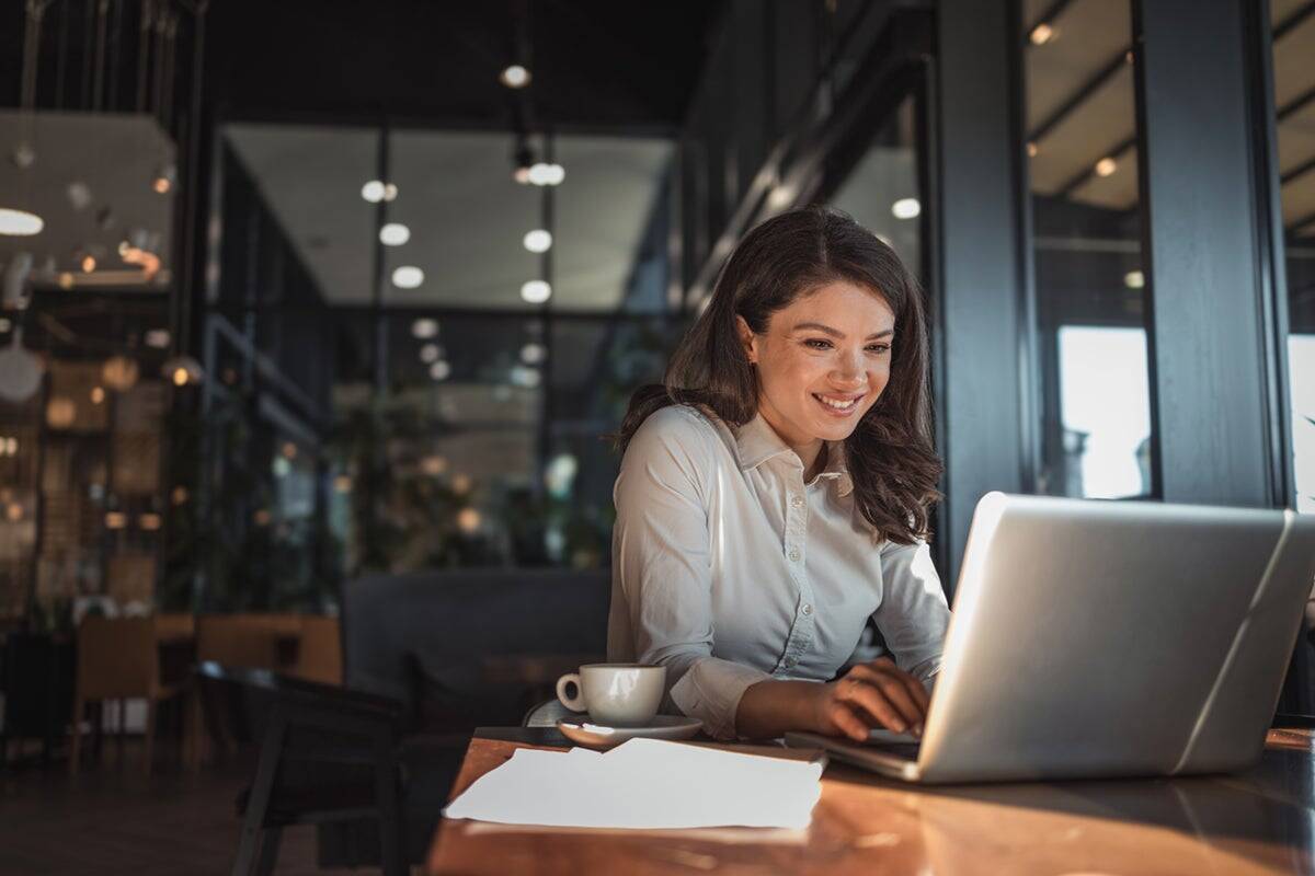 Woman smiling while working on a laptop in a modern café, with a coffee cup and papers on the table.