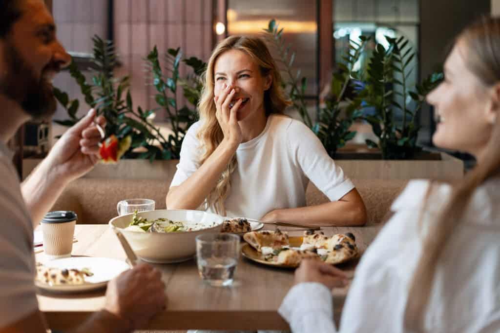 Three people laughing and eating together at a restaurant table with salads and pizza.
