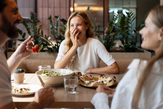 Three people laughing and eating together at a restaurant table with salads and pizza.
