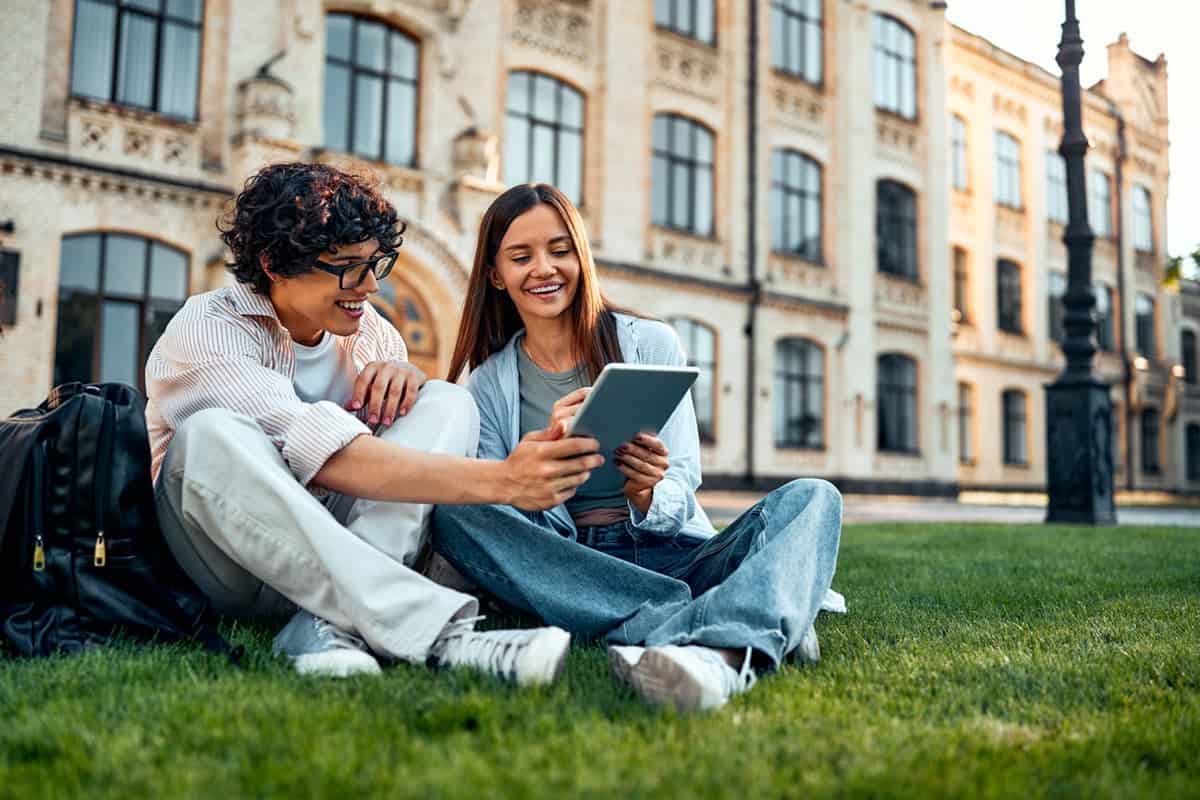 Two students sitting on grass outside, smiling and looking at a tablet together in front of a building.