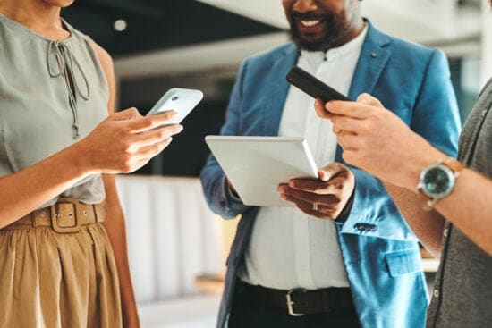 Three people using smartphones and a tablet, standing together and smiling in a modern office setting.