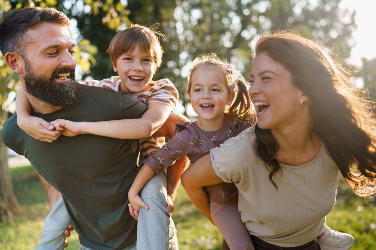 Two smiling adults give piggyback rides to two laughing children outdoors on a sunny day.