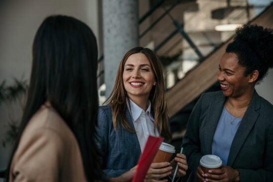 Three women in business attire smiling and talking while holding coffee cups in an office setting.