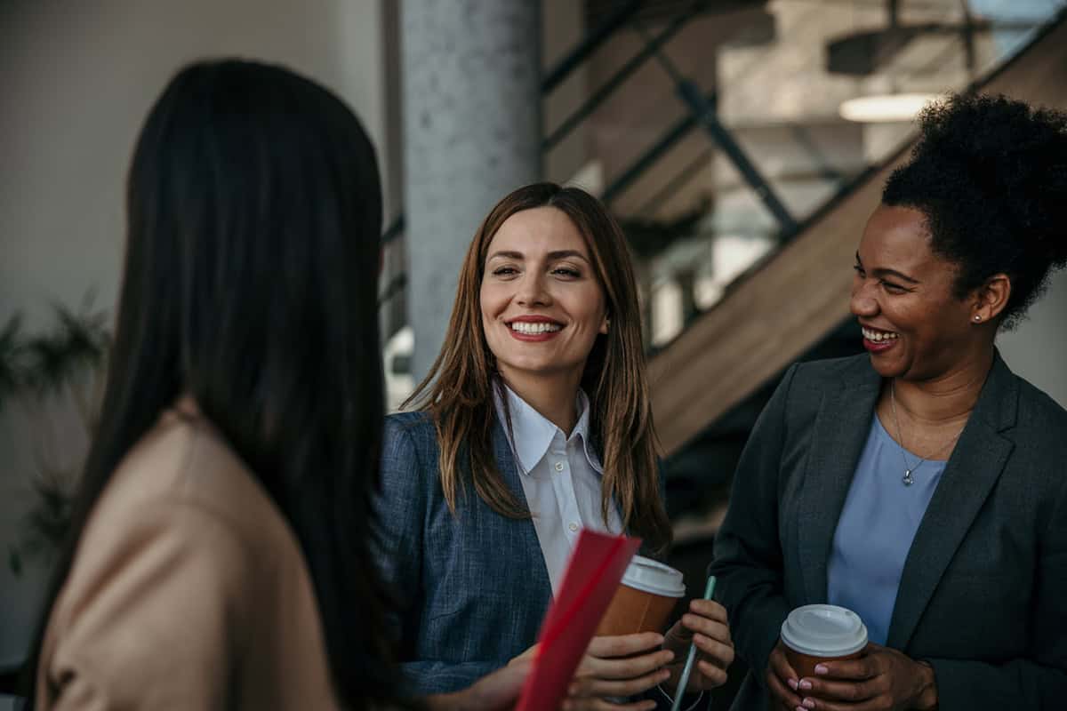 Three women in business attire smiling and talking while holding coffee cups in an office setting.