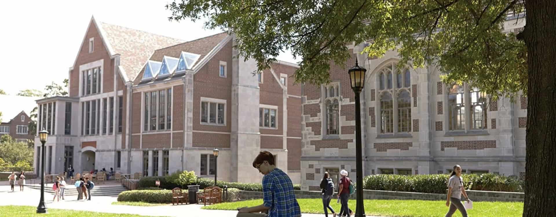 Students walk and relax outside a large brick and stone campus building on a sunny day.