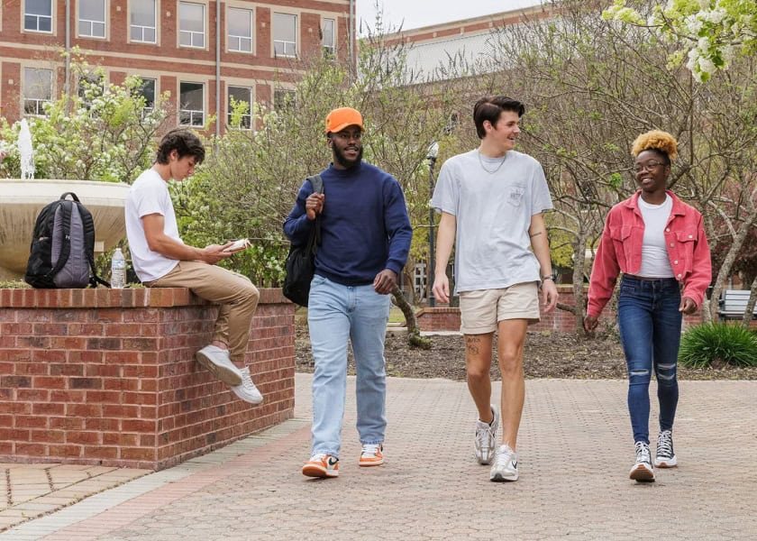 Four college students walk and chat outside on campus; one sits on a brick wall looking at a notebook.