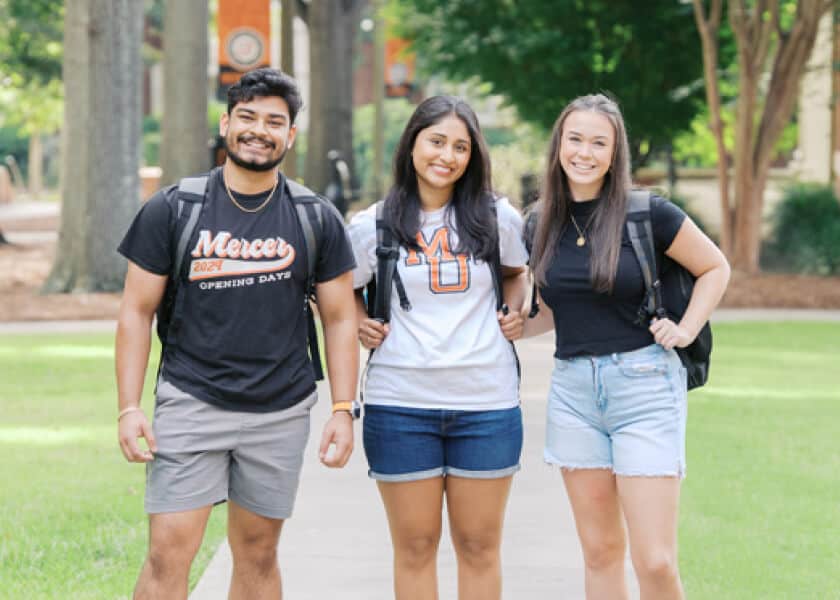 Three students with backpacks smile while standing on a campus walkway surrounded by trees.
