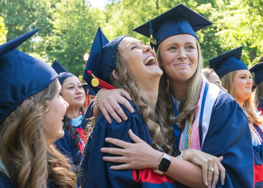 Two graduates in blue caps and gowns hug and smile outdoors during a graduation ceremony.