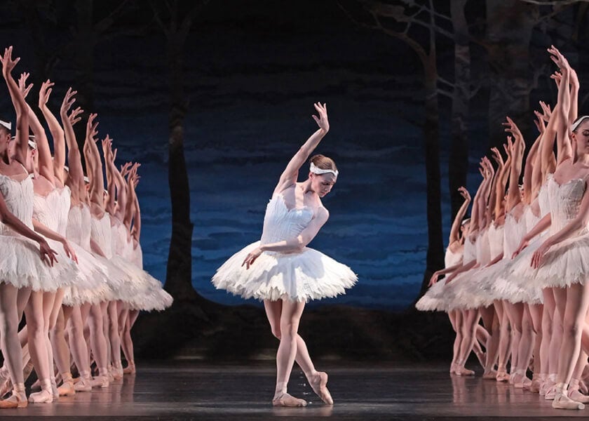 Ballet dancers in white tutus perform on stage, with the central dancer posing gracefully under blue lighting.