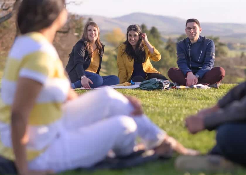 Four people sit on grass in a park, smiling and talking, with hills in the background.