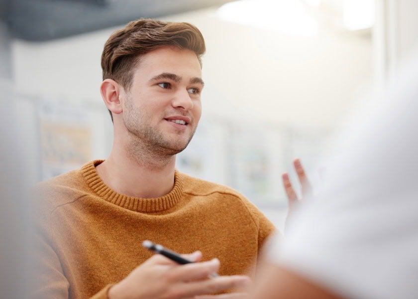 Young man in a brown sweater talking, holding a pen, gesturing with his hand in a bright indoor setting.