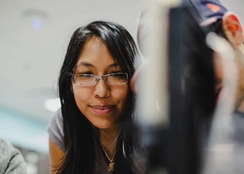 Woman with glasses smiling and looking at something off-camera; blurred background and foreground.