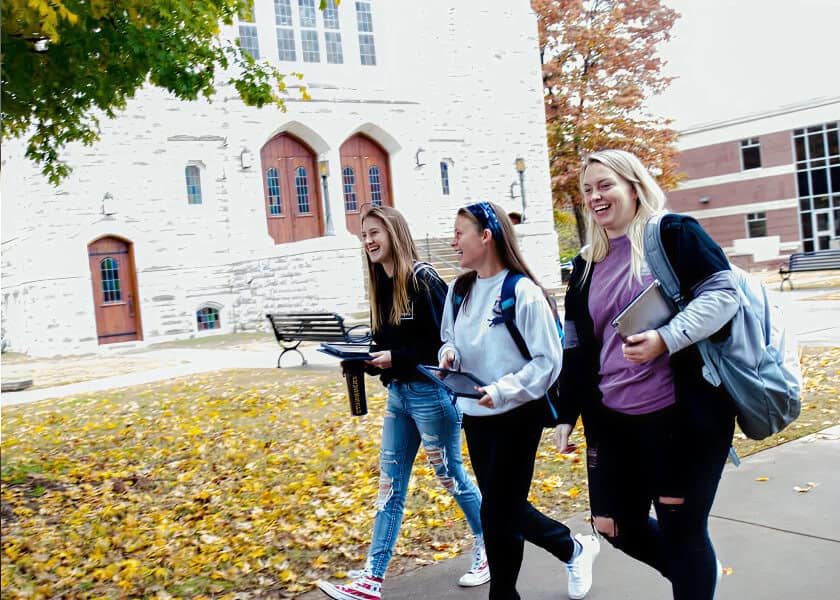 Three smiling students walk across a campus sidewalk with books and backpacks on a fall day.