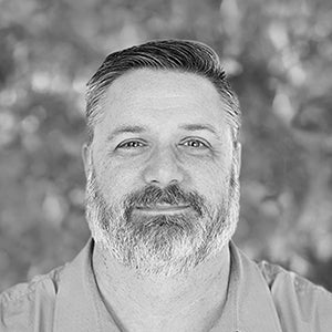 Man with short hair and a beard, wearing a collared shirt, looking at the camera, blurred background.