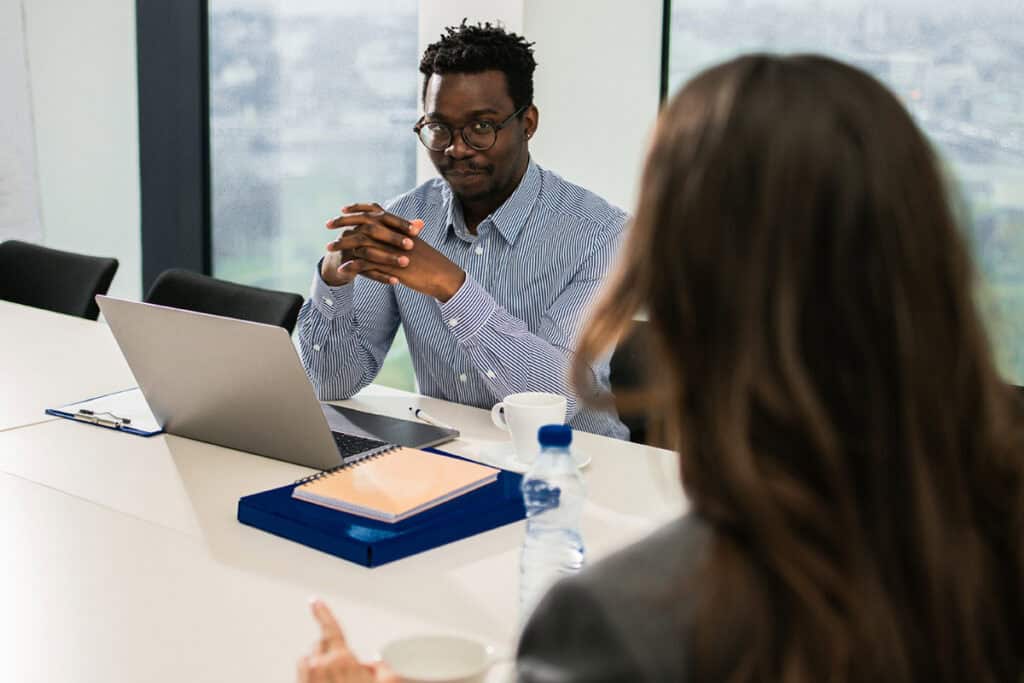 Man in glasses sits at a desk with a laptop, listening to a woman during a business meeting.