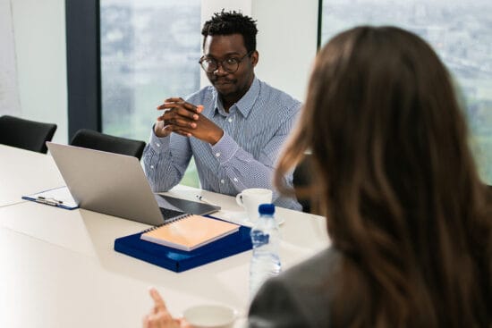 Man in glasses sits at a desk with a laptop, listening to a woman during a business meeting.