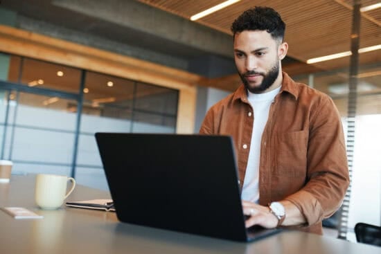 Man in a brown shirt working on a laptop at a desk with a mug and notebook nearby.