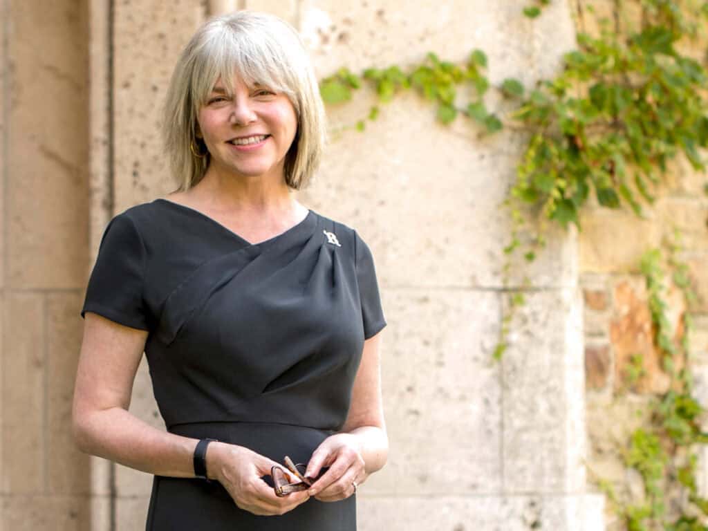 Smiling woman with gray hair in a black dress stands in front of a stone wall with green ivy.