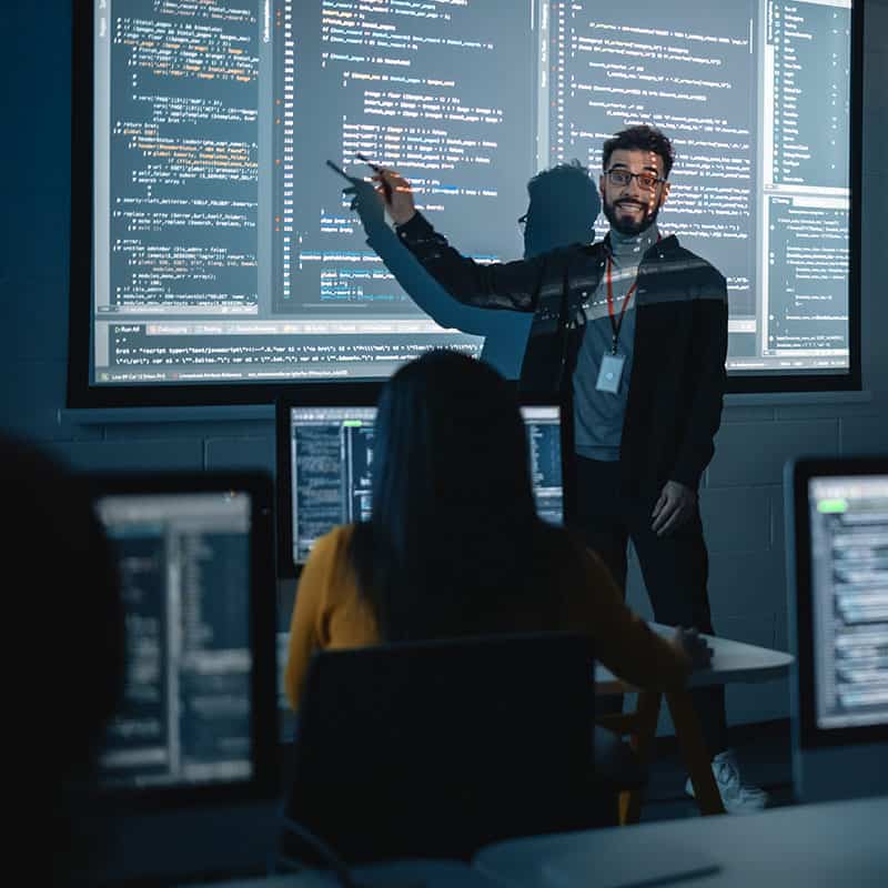 Man pointing at projection with code on it in front of class.