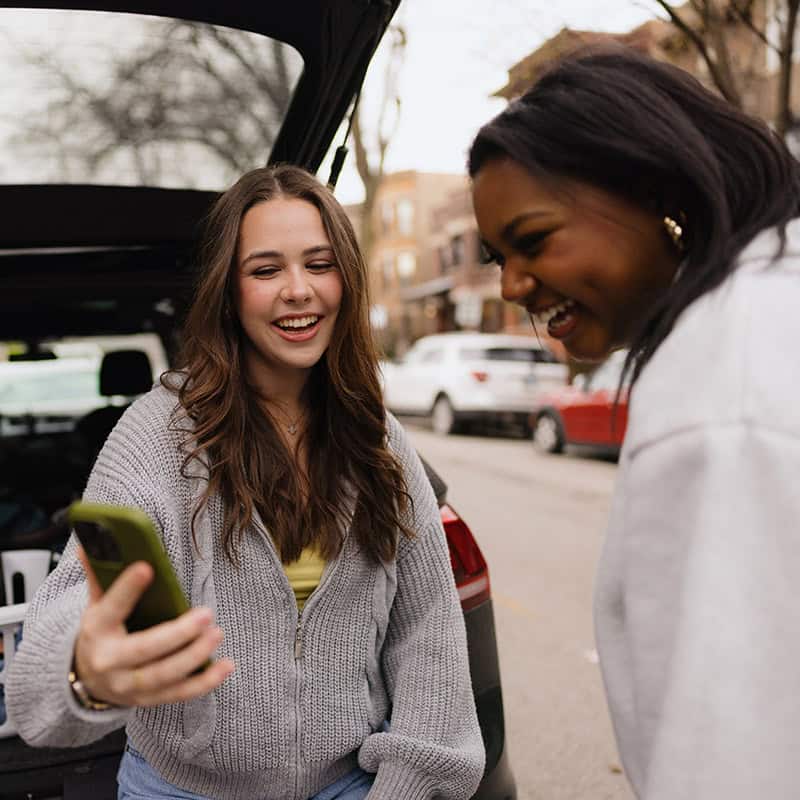 Two women outside of car video chatting with someone else on a smart phone