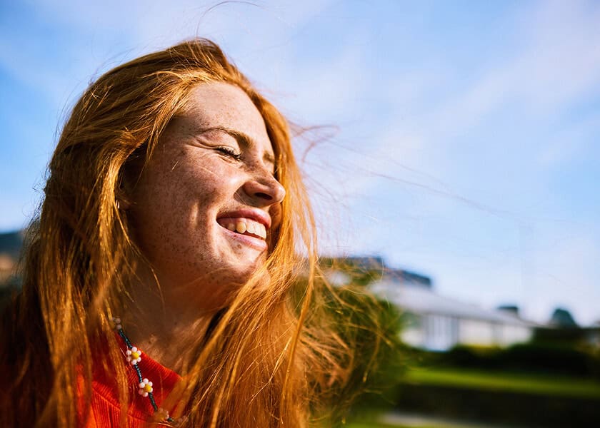 Smiling woman with long red hair enjoying sunlight outdoors, eyes closed, wind blowing her hair.