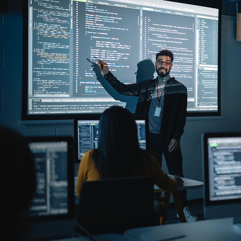 Man pointing at projection with code on it in front of class.