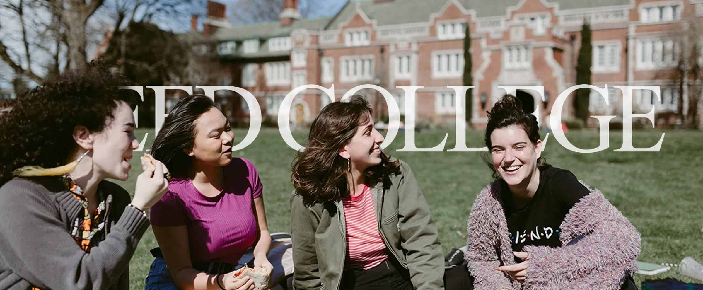Four young women sit on grass, smiling and talking, with a large brick college building in the background.