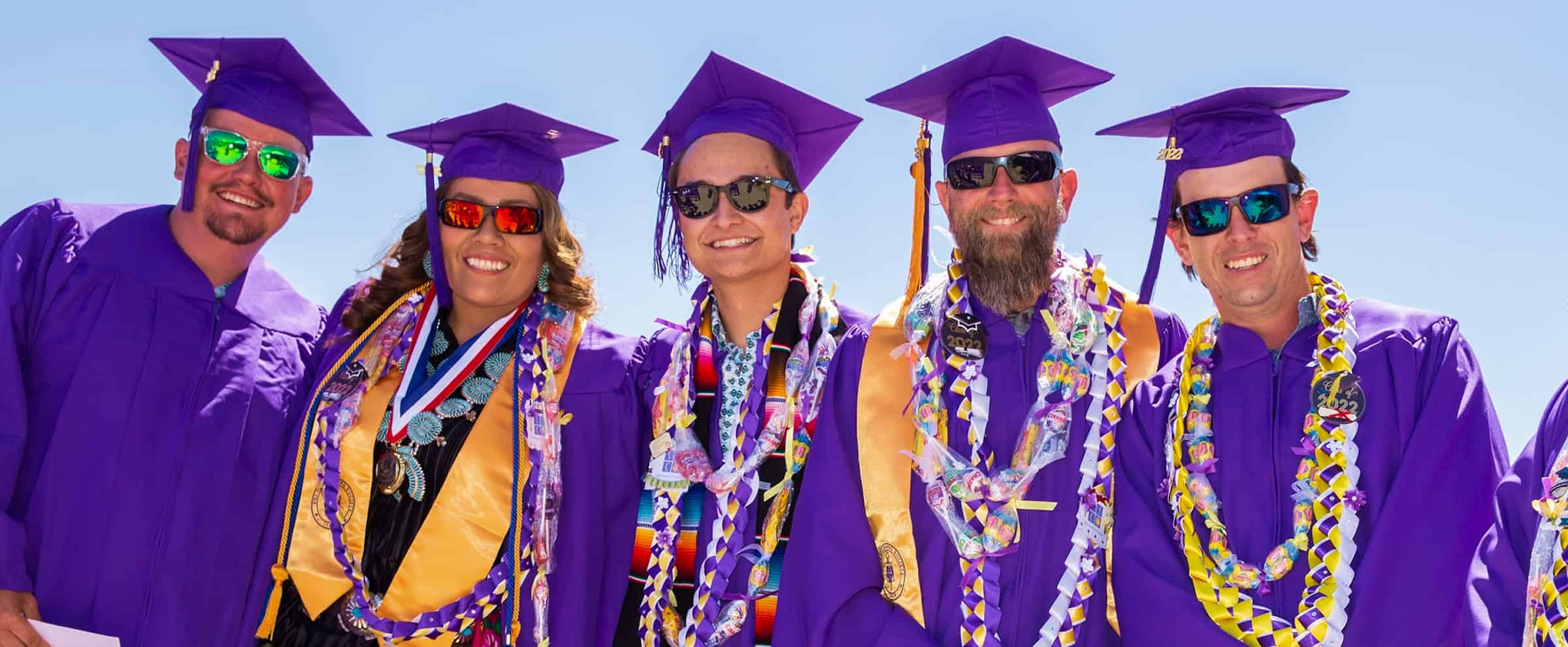 Five graduates in purple gowns and caps, smiling and wearing leis and sunglasses, stand together outdoors.