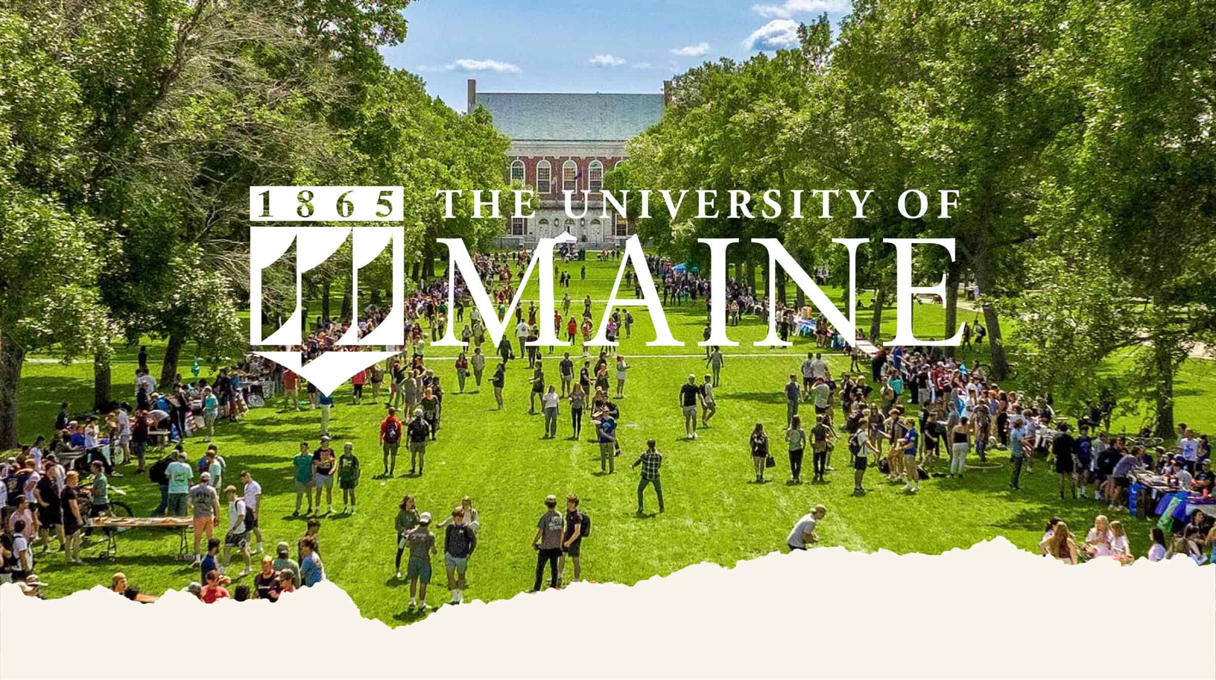 A large group of people gathers on a green lawn in front of a campus building at the University of Maine.