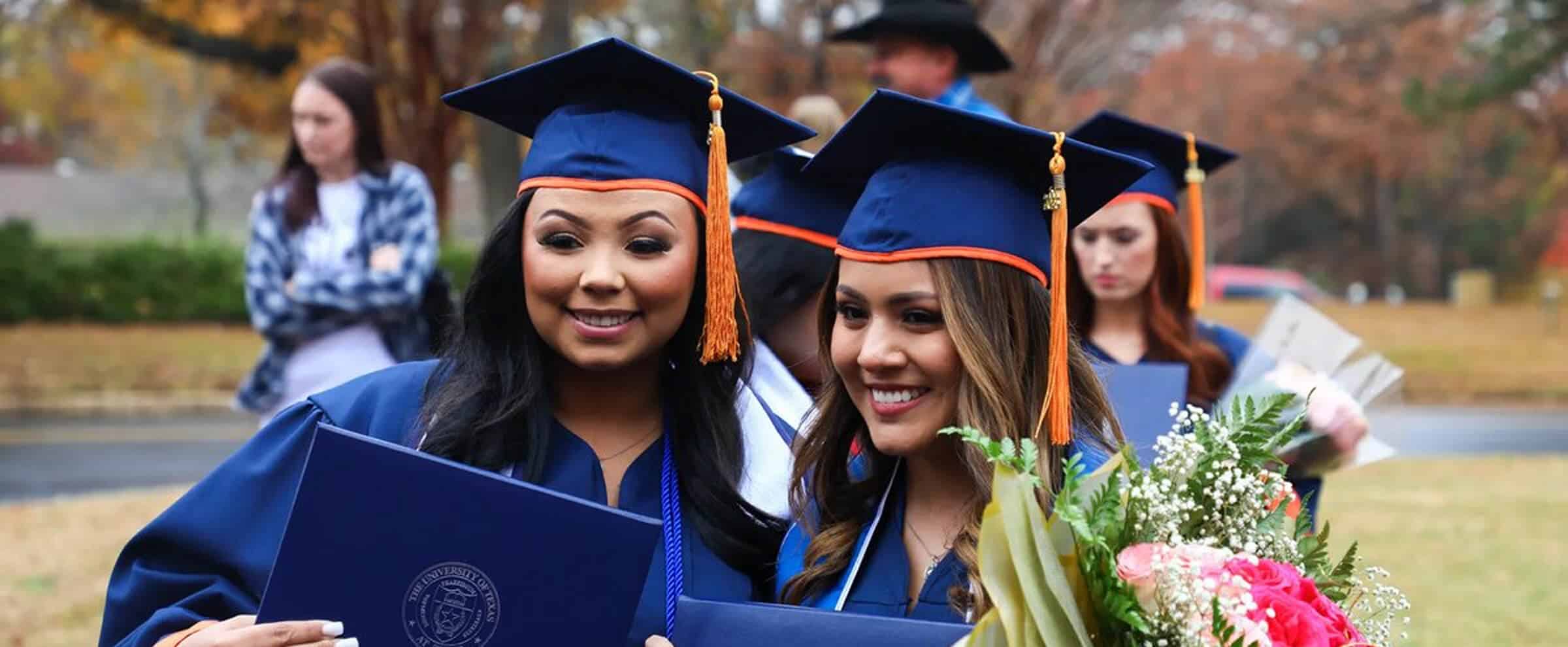 Two graduates in caps and gowns smile, holding diplomas and flowers at an outdoor graduation ceremony.