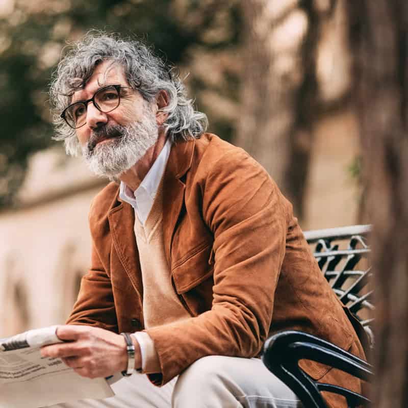 Older man with gray hair and glasses sits on a bench outdoors, holding a newspaper and looking thoughtful.