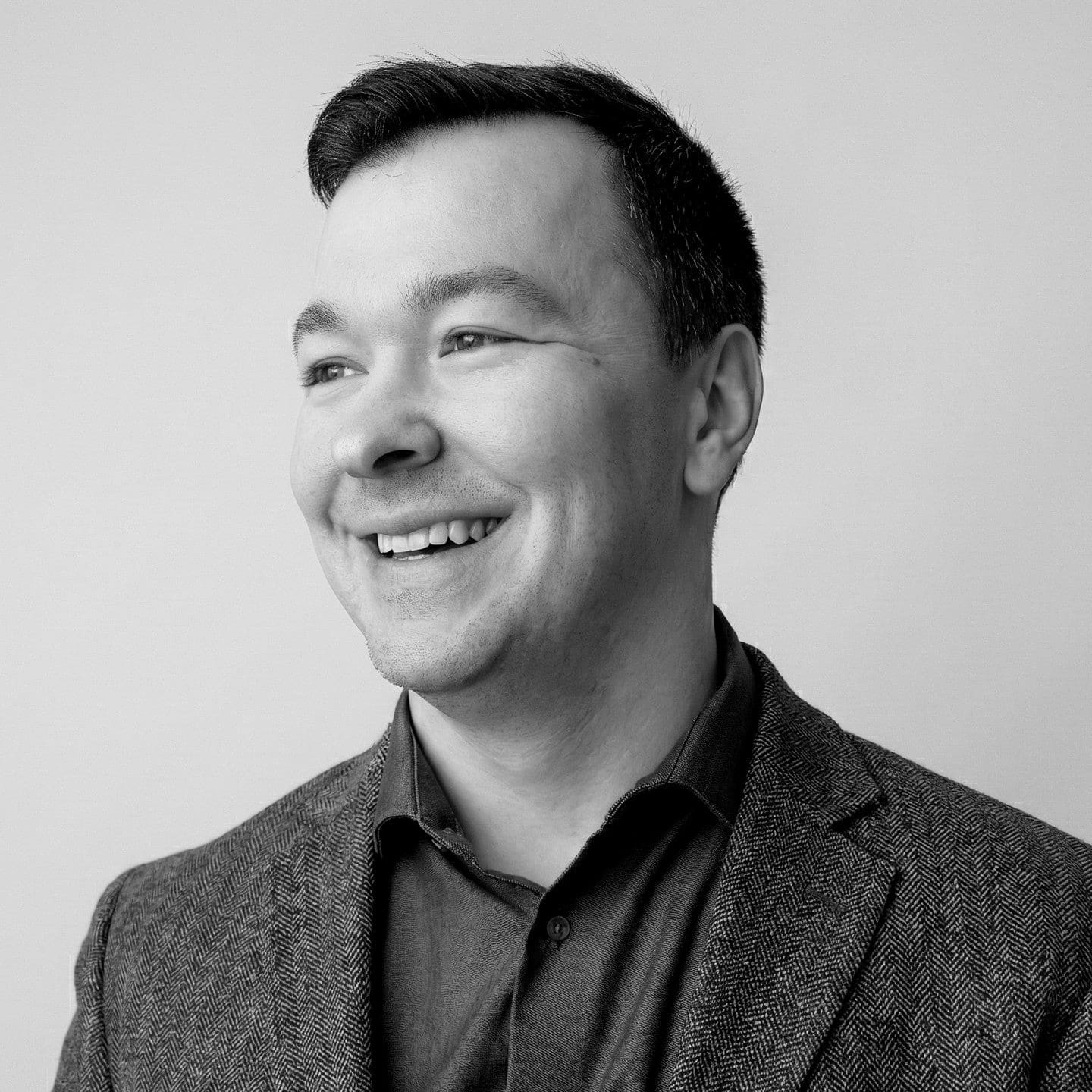 Man in a suit jacket smiling and looking to the side against a plain background.