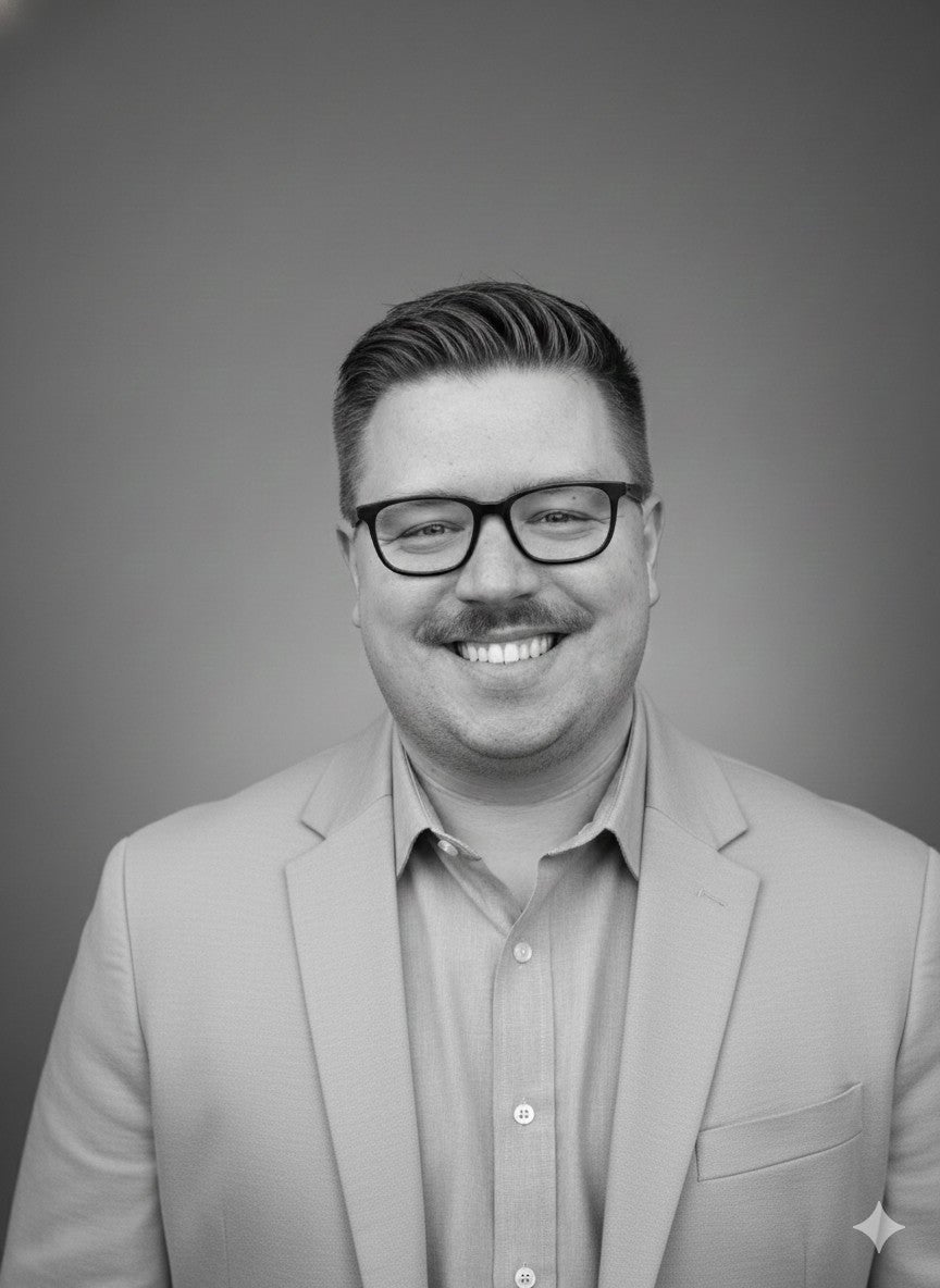 Smiling man with glasses and a mustache in a light blazer, posing against a plain background.