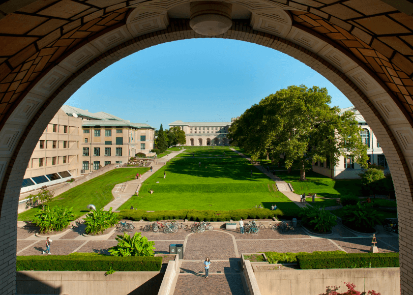 View of a large grassy quad and campus buildings framed by an archway on a bright, sunny day.