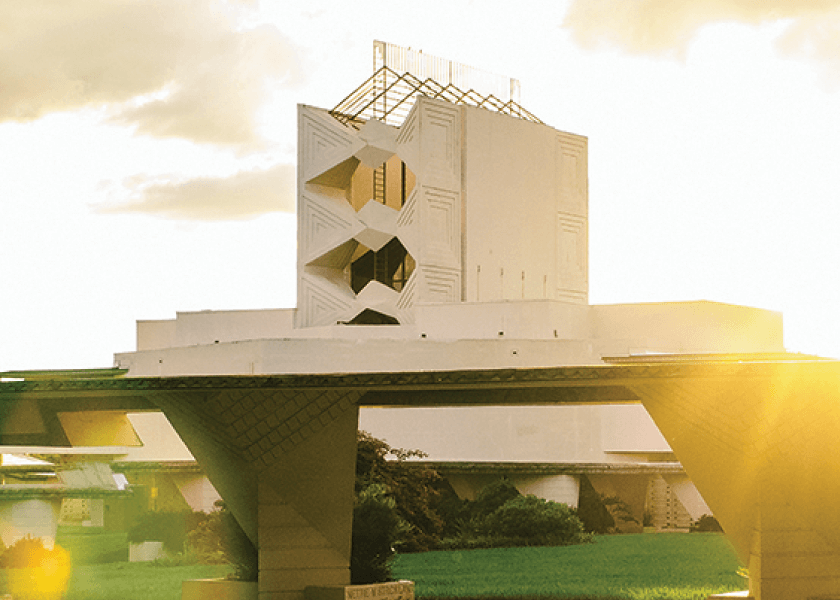 Modern white building with geometric designs, surrounded by greenery in warm, golden sunlight.