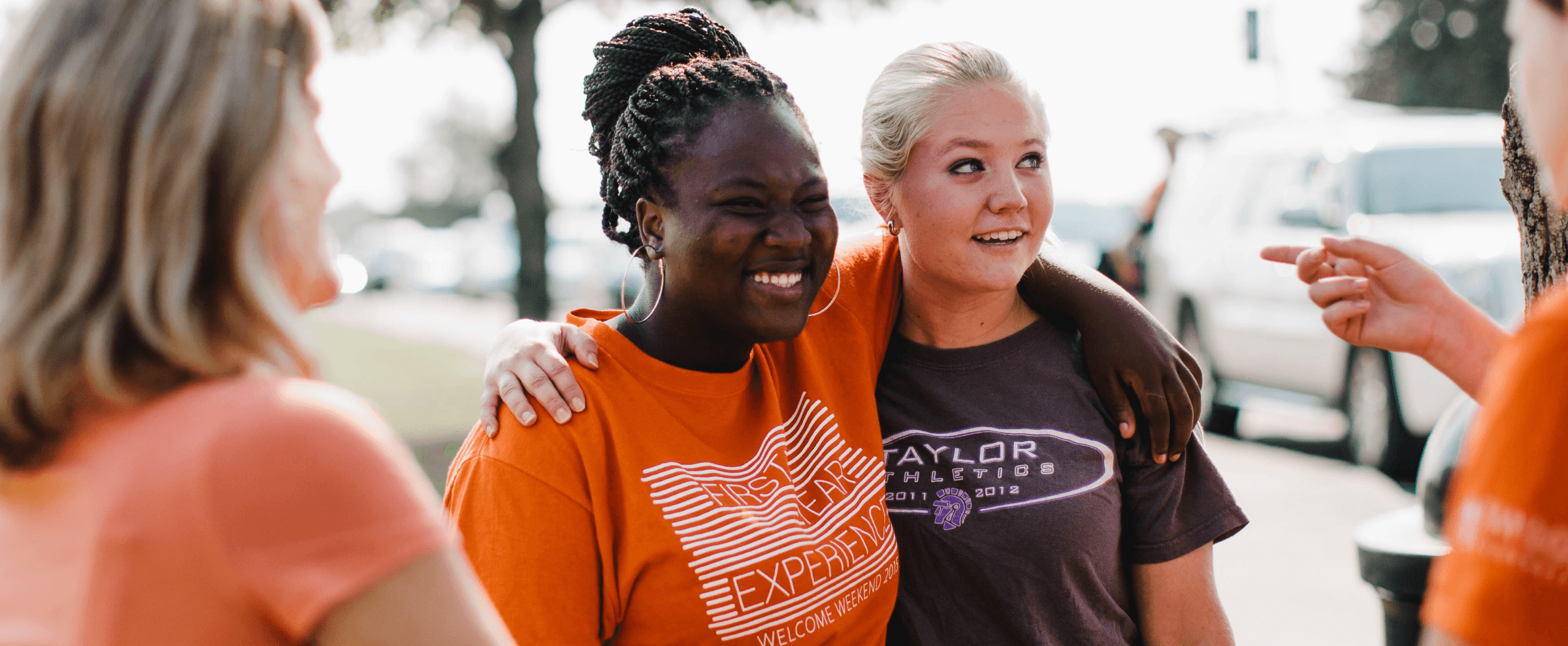 Two young women smile and hug outdoors while friends socialize around them on a sunny day.