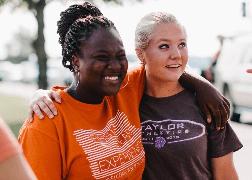 Two young women smiling with arms around each other outdoors, wearing casual T-shirts.