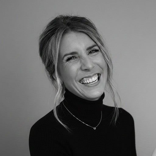 Woman with light hair smiling, wearing a black turtleneck and necklace, in front of a plain background.