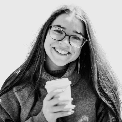 Young woman with glasses smiling and holding a coffee cup, wearing a sweater, in a black and white photo.