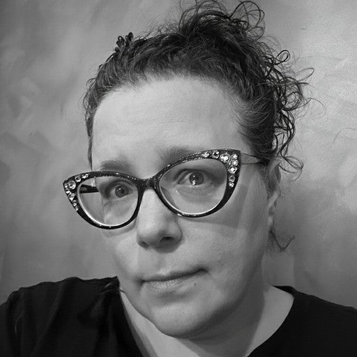 Woman with curly hair and embellished glasses looks at the camera, wearing a black shirt, grayscale image.