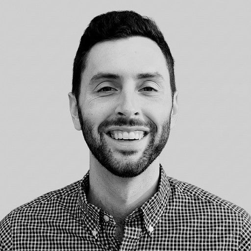 Smiling man with a beard, wearing a checkered shirt, standing against a plain background.