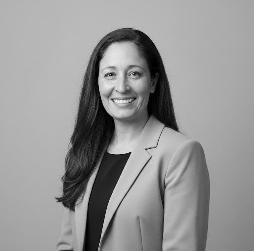 Woman with long dark hair in a light blazer, smiling, posed against a plain background in a black-and-white photo.