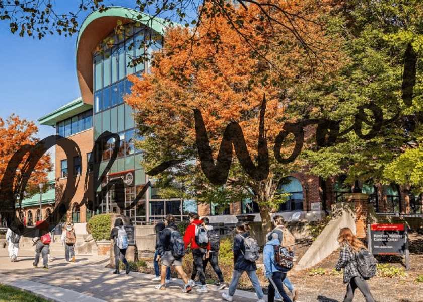 Students walk on a campus path with autumn trees and a modern brick building in the background.